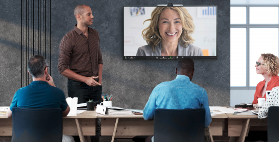 Three people sit around a table while a man stands to present to a woman on a RingCentral video call